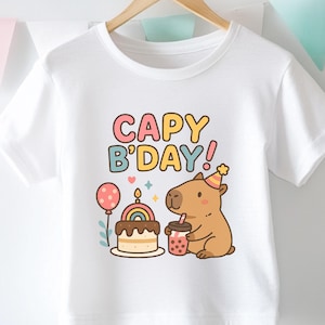 May include: White t-shirt with a colorful graphic that says "CAPY B'DAY!" featuring a cartoon capybara wearing a party hat, a cake, a rainbow, and a drink. The shirt is hanging on a wooden hanger.