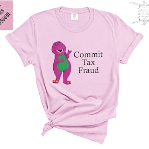 May include: A light pink short-sleeve t-shirt featuring a graphic of a purple dinosaur with a green belly and the black text "Commit Tax Fraud" printed to the right. The shirt has a crew neckline and a small tag visible at the inner collar area.