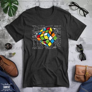 May include: A dark gray short-sleeve t-shirt featuring a graphic of a colorful Rubik's cube surrounded by white mathematical equations and formulas. The cube displays red, blue, yellow, green, and white squares with white circular arrows.