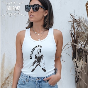 May include: A white ribbed tank top featuring a black graphic print of a musician holding a guitar with the text SPRINGSTEEN arched above the figure. The garment has a scoop neckline and is paired with light blue denim jeans.