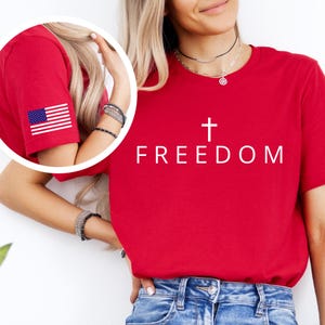 May include: A red t-shirt with the word "FREEDOM" and a white cross graphic. The left sleeve has a small American flag. The shirt is worn with blue denim jeans.