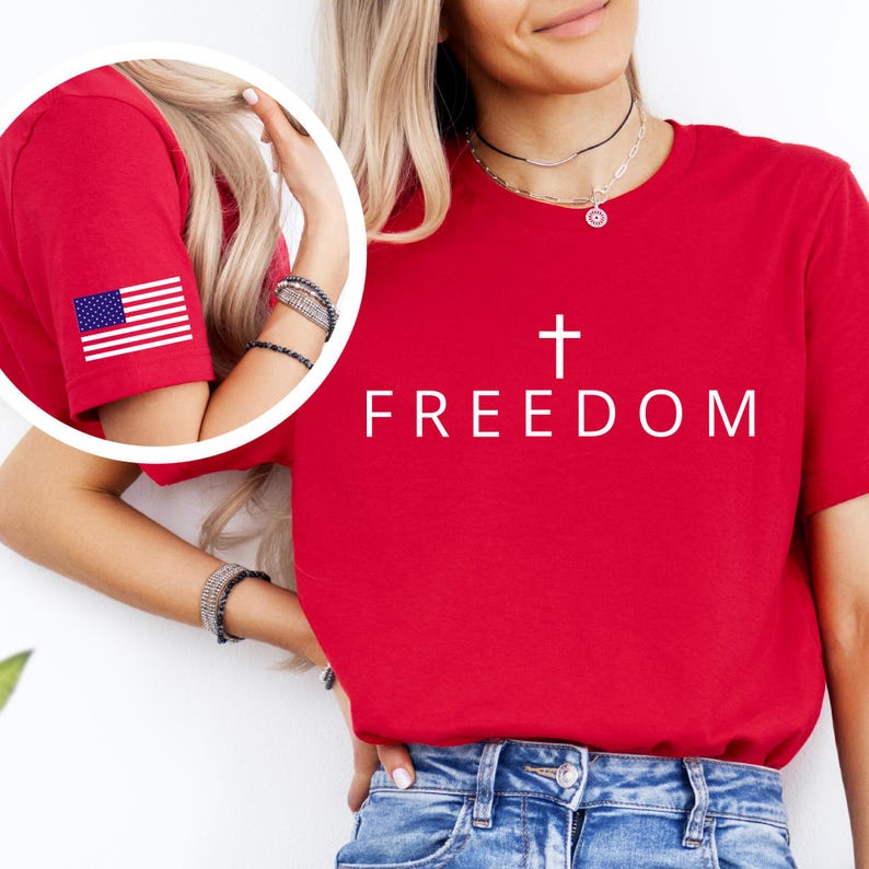 May include: A red t-shirt with the word "FREEDOM" and a white cross graphic. The left sleeve has a small American flag. The shirt is worn with blue denim jeans.