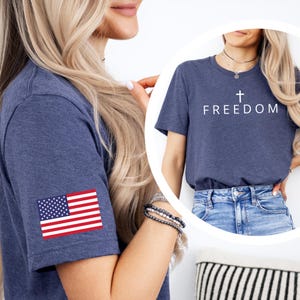 May include: Navy blue t-shirt with the word "FREEDOM" and a cross printed in white. The shirt also features a small American flag patch on the sleeve. The shirt is made of a soft material.