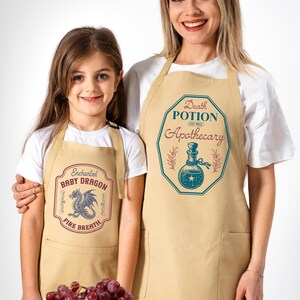 May include: Two beige aprons with graphic designs. One apron features a dragon and the text "Enchanted Baby Dragon Fire Breath." The other apron has a potion bottle and reads "Death Potion Apothecary." Both aprons are worn by a woman and a child.