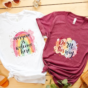 May include: Two t-shirts with graphic designs. One is white with a rainbow and the text "everyone is welcome here." The other is maroon with the text "born this way." Both shirts are short-sleeved.