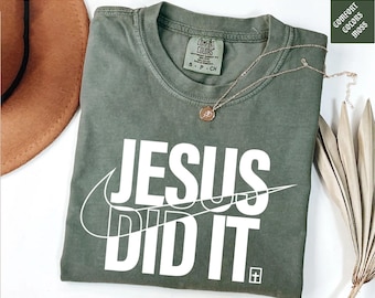 May include: A moss green short-sleeved t-shirt featuring the white text JESUS DID IT with a swoosh graphic and a small cross icon. The shirt has a Comfort Colors brand label at the inner neckline indicating size S and 100% cotton material composition.