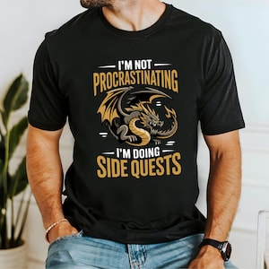 May include: A black short-sleeved t-shirt featuring a graphic of a gold and grey dragon in the center. The text above the dragon reads I'M NOT PROCRASTINATING and the text below the dragon reads I'M DOING SIDE QUESTS in bold gold lettering.