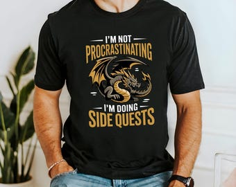 Peut inclure: Un t-shirt noir à manches courtes avec un motif central représentant un dragon doré et gris. Le texte au-dessus du dragon indique I'M NOT PROCRASTINATING et le texte en dessous indique I'M DOING SIDE QUESTS en lettres dorées grasses.