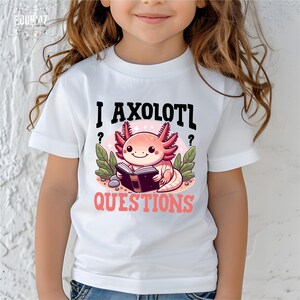 May include: A white short-sleeved t-shirt featuring a graphic of a pink axolotl reading a book. The text above reads I AXOLOTL and the text below reads QUESTIONS in pink. The design includes green leaves and small gray stones on a light pink circle.