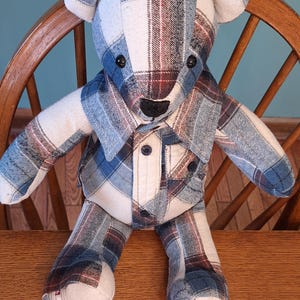 May include: A plaid teddy bear made from repurposed fabric. The bear is sitting on a wooden chair. The bear has a white, blue, and red plaid pattern.