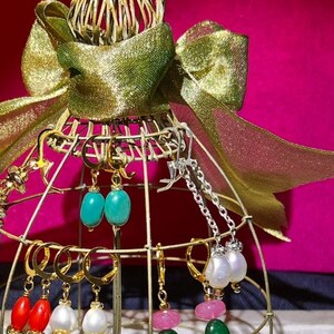 May include: A gold metal display stand with a gold bow on top, holding a variety of earrings with turquoise, white pearl, red, and green stones.