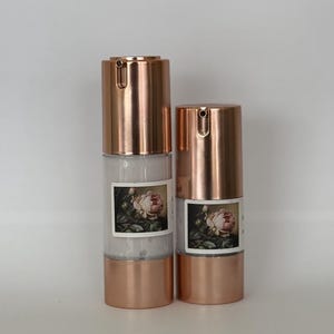 May include: Two rose-gold bottles with a white label featuring a vintage floral illustration. The bottles have a pump dispenser and are filled with a white liquid.