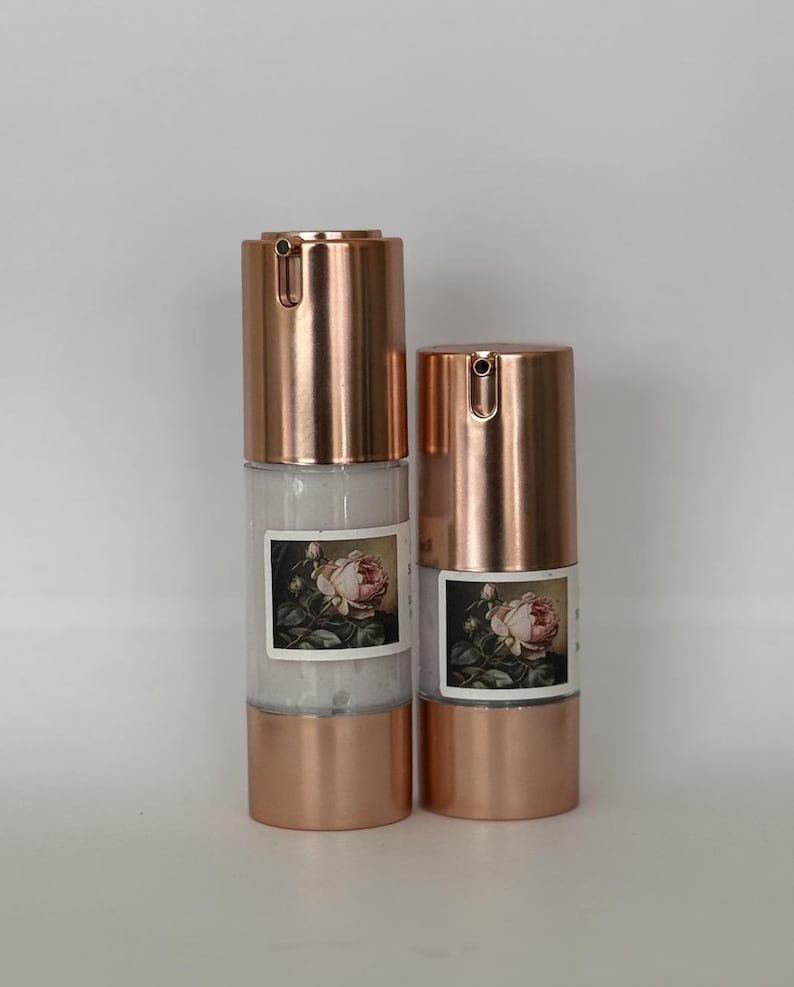 May include: Two rose-gold bottles with a white label featuring a vintage floral illustration. The bottles have a pump dispenser and are filled with a white liquid.