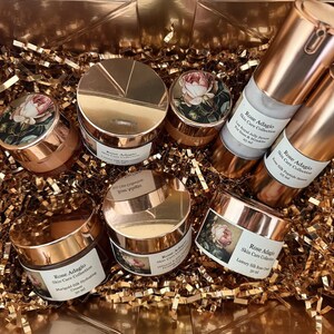 May include: A set of six rose-gold coloured jars and bottles with labels that read "Rose Adagio Skin Care Collection". The jars and bottles are filled with a white cream and a clear liquid. The jars and bottles are arranged in a basket filled with gold-coloured tissue paper.
