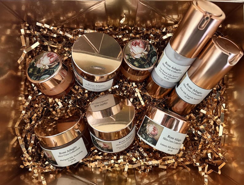 May include: A set of six rose-gold coloured jars and bottles with labels that read "Rose Adagio Skin Care Collection". The jars and bottles are filled with a white cream and a clear liquid. The jars and bottles are arranged in a basket filled with gold-coloured tissue paper.