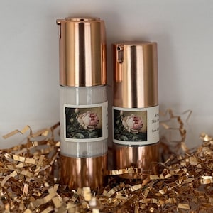 May include: Two rose-gold bottles of rose-scented skincare products with a rose graphic on the label. The bottles are sitting on a bed of gold tissue paper.