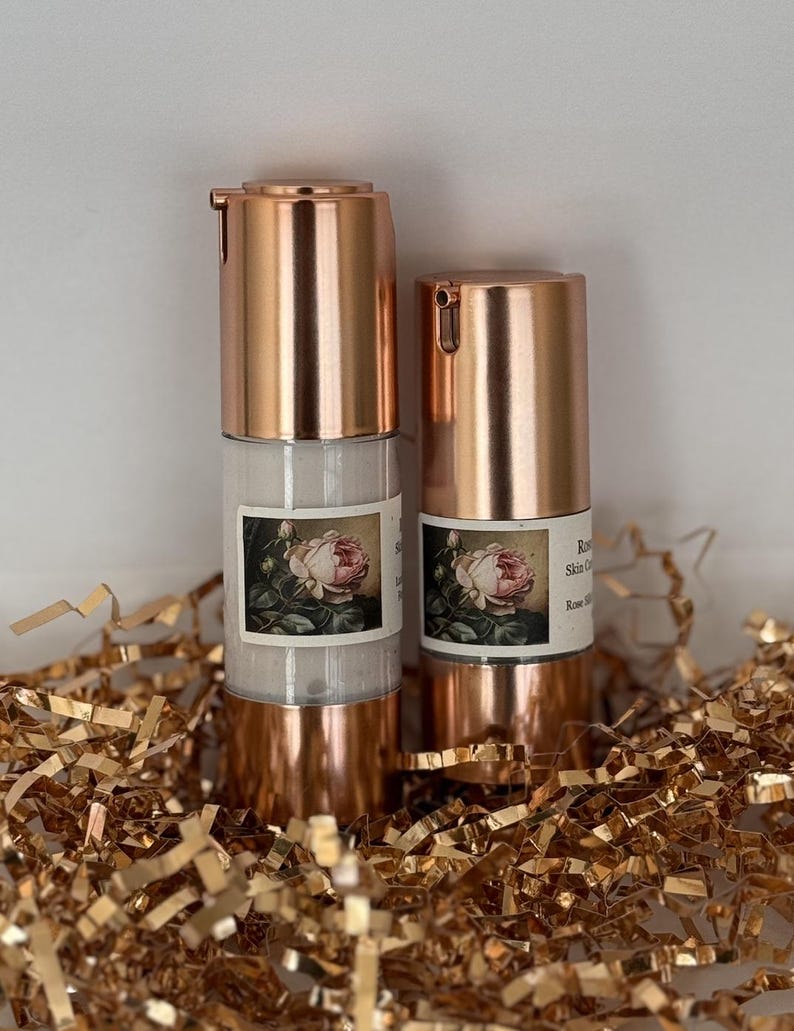 May include: Two rose-gold bottles of rose-scented skincare products with a rose graphic on the label. The bottles are sitting on a bed of gold tissue paper.
