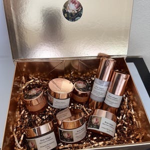 May include: A set of six rose-themed skincare products in a gold gift box. The products include a face cream, a serum, and a lotion. The box is filled with gold tissue paper.