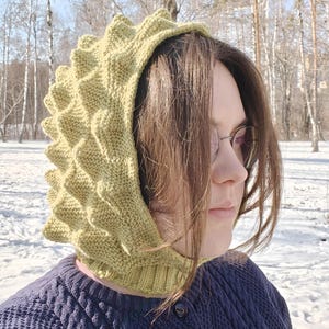May include: A person wearing a green knitted hooded hat with a textured, spiky design. The hat is pulled down over their head and covers their ears. The person is wearing a blue sweater and is standing in a snowy forest.