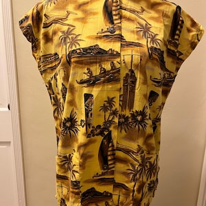 Può includere: Camicia hawaiana gialla con stampa tropicale marrone con aerei, barche, palme e statue tiki. La camicia ha un colletto alla coreana e maniche corte. Il tessuto ha un aspetto leggermente strutturato.