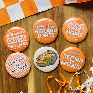 May include: A collection of orange and white round buttons with text, including "Straight Outta Neyland," "Under those Neyland Lights," and "Home Sweet Home to Me." One button features a stadium illustration.