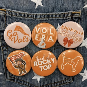 May include: A set of six circular pin-back buttons featuring orange and white designs. Text includes Go Vols, Vol Era, Knoxville Saturdays, Good ol Rocky Top, and Good Boy. Graphics include a mascot, a bow, white boots, and a dog silhouette.