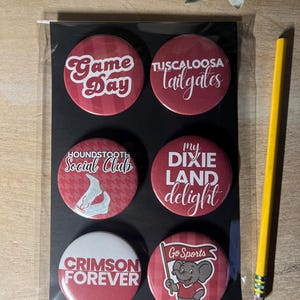 May include: A set of six round, red and white buttons with various text and graphics. The buttons include phrases like "Game Day," "Tuscaloosa Tailgates," and "Crimson Forever." A yellow pencil rests beside the buttons.
