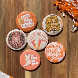 May include: A collection of six round, orange and white Dolly Parton-themed buttons. The buttons feature images of Dolly, text like "Raised on Dolly & UT Football," and phrases like "Big Dolly Energy" and "What Would Dolly Do?"