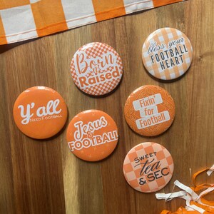 May include: A collection of orange and white football-themed buttons with various phrases, including "Born & Raised," "Y'all Need Football," and "Sweet Tea & SEC." The buttons are arranged on a wooden surface with a checkered orange and white fabric.