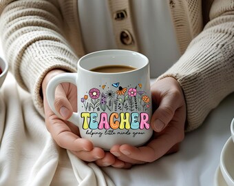 May include: White ceramic mug with a colorful floral design and the word "TEACHER" in rainbow letters. The mug also has the text "helping little minds grow". The mug is filled with a dark liquid.