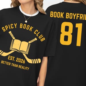 Op de afbeelding: Zwart T-shirt met witte kraag met een gouden opdruk. De voorkant toont "SPICY BOOK CLUB" met gekruiste hockeysticks en een boek, "EST. 2026 BETTER THAN REALITY." De achterkant leest "BOOK BOYFRIEND 81" in goud.