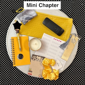 May include: A collection of stationery items, including a yellow spiral notebook, pen, candle, bookmark, and scrunchies. The items are arranged on a white plate with a black and white polka dot background. The text "Mini Chapter" is at the top.