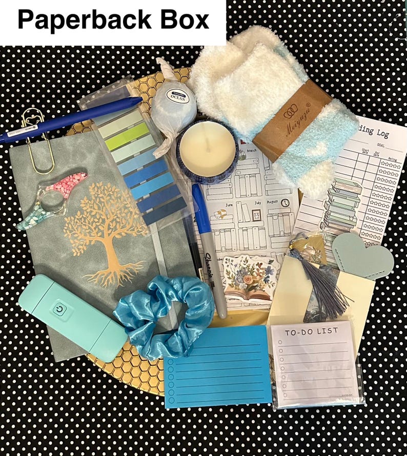 May include: A curated gift box with a variety of items. Includes a journal, pen, candle, scrunchie, notepad, and other stationery items. The box is on a black and white polka dot background. The text "Paperback Box" is at the top.