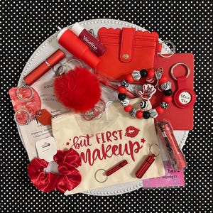May include: A collection of red accessories, including a wallet, keychain, lip balm, and a small bag with the text "but FIRST makeup". The items are arranged on a white plate against a black and white polka dot background.