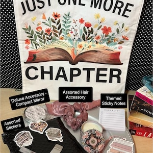 May include: A white tote bag with the text "JUST ONE MORE CHAPTER" and a floral design. The bag contains a compact mirror, hair accessory, stickers, sticky notes, pen, pampering item, and a bookmark. The items are on a silver tray.