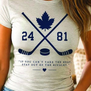 May include: A light gray heathered t-shirt featuring a dark blue graphic of two crossed hockey sticks, a maple leaf, a hockey puck, and the numbers 24 and 81. Below is the text: IF YOU CAN'T TAKE THE HEAT, STAY OUT OF THE RIVALRY. with a heart icon.