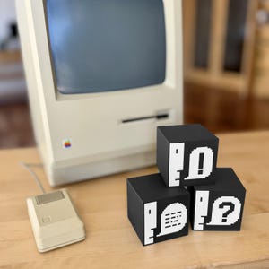 May include: A vintage beige computer with a gray screen, a matching mouse, and three black cubes with white pixelated graphics. The cubes display an exclamation point, a zero, and a question mark.