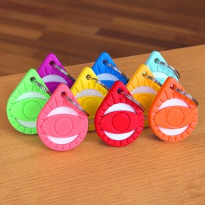 May include: A collection of eight teardrop-shaped keychains in various colors including red, orange, yellow, green, blue, purple, and pink. Each keychain features a central eye-shaped design with a white background and a metal ring attached.