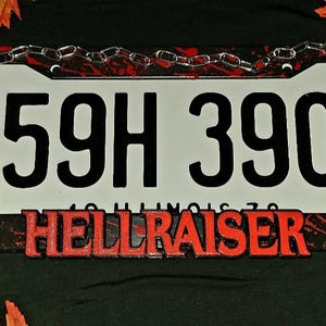 May include: A red, white, and black license plate with the number 59H 390 and the words "Hellraiser" printed on it. A small, metal box with a geometric design is on the right side of the plate. A small, white, spiky head with a face is on the left side of the plate.