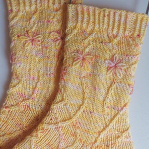 May include: A pair of yellow knitted socks with a white and pink flecked yarn. The socks have a cable knit pattern and embroidered flowers.