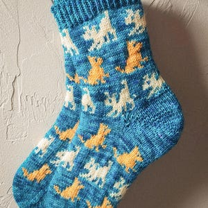 Puede incluir: Un par de calcetines azules de punto con un patrón de gatos blancos y naranjas. Los calcetines están colgados en una pared blanca.