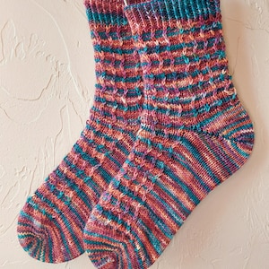 Puede incluir: Un par de calcetines de punto coloridos con un patrón de diamantes texturizado. Los calcetines presentan una mezcla de tonos rojos, naranjas, turquesas y morados. Los calcetines están colgados de un alambre metálico.