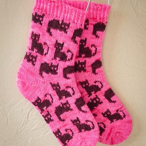 以下が含まれることがあります： 茶色の猫の模様がついたピンクのニットソックスのペア。ソックスは金属製のハンガーにかけられています。