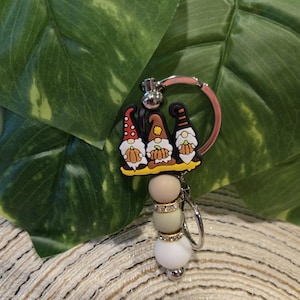 May include: A keychain featuring three gnome figures with red, brown, and black hats. The keychain has a pink ring, silver hardware, and a beaded charm with beige, green, and white beads. The beads are accented with rhinestones.