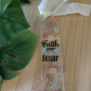 Op de afbeelding: Transparante acryl bladwijzer met de tekst "Faith over fear". De bladwijzer heeft kleurrijke illustraties van een regenboog, cactus en potplant. Bovenop is een crèmekleurig lint bevestigd.