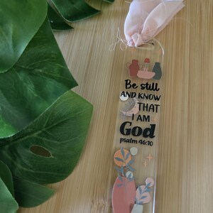 Peut inclure: Marque-page en acrylique transparent avec un ruban rose et le texte "Be still AND KNOW THAT I AM God psalm 46:10." Le marque-page présente des illustrations de vases et de plantes aux couleurs douces.