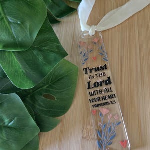 May include: A clear acrylic bookmark with the text "Trust in the Lord with all your heart Proverbs 3:5". The bookmark features pink flowers, blue leaves, and red hearts. A white ribbon is attached at the top. The bookmark is on a wooden surface.