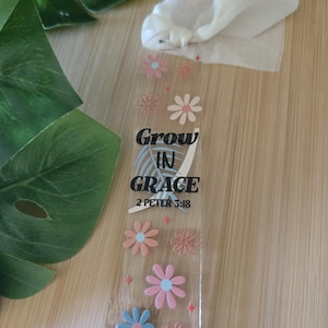 Peut inclure: Marque-page transparent avec l'inscription "Grow in Grace 2 Peter 3:18" et des motifs floraux roses et bleus. Un ruban blanc est attaché. Le marque-page est posé sur une surface en bois.