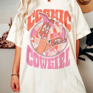 May include: A white t-shirt with a pink graphic design featuring a pair of pink cowboy boots and the text "COSMIC COWGIRL".