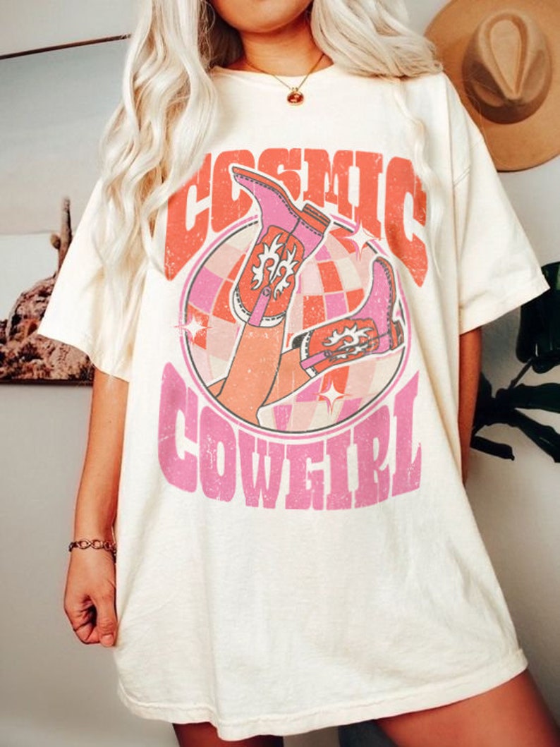 May include: A white t-shirt with a pink graphic design featuring a pair of pink cowboy boots and the text "COSMIC COWGIRL".